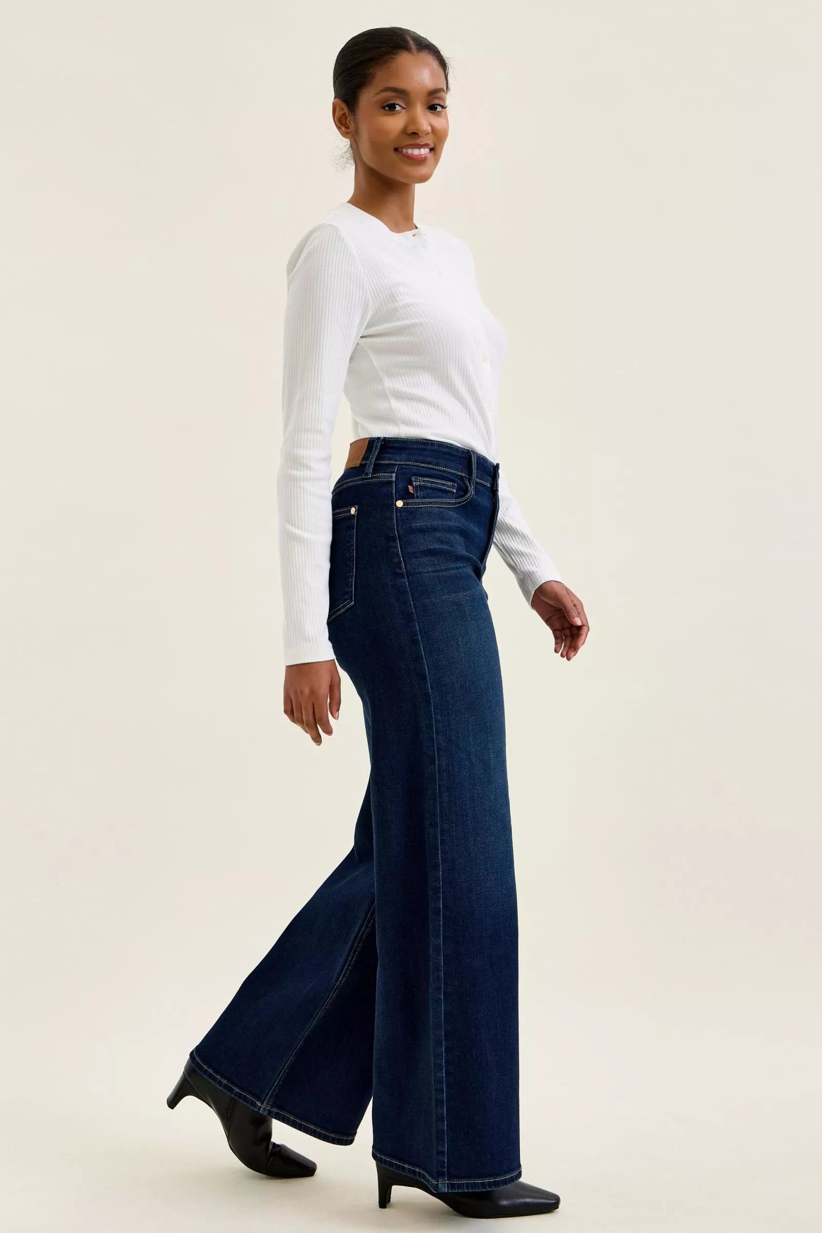 Model wearing dark blue high-waisted wide-leg jeans with a white long-sleeve top and black heels, showcasing a stylish look.