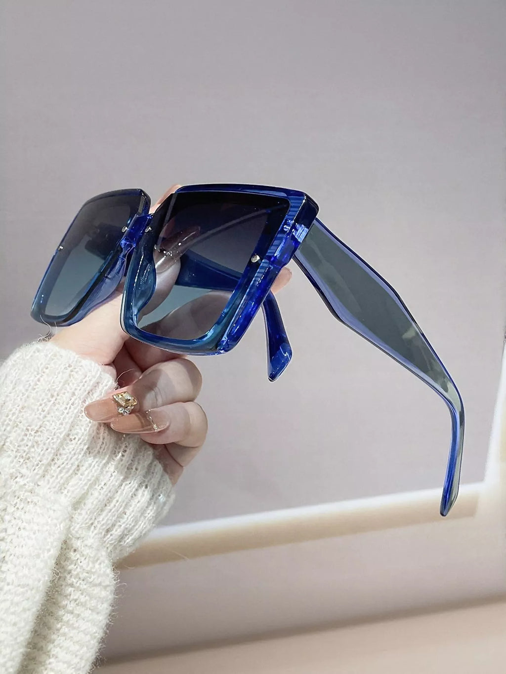 Hand holding oversized square sunglasses with blue frame and UV400 lenses against a neutral background.