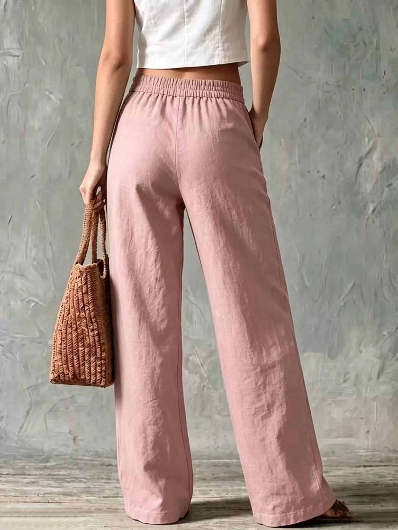 Back view of a woman wearing plus size wide leg pants in soft pink, paired with a white crop top and a woven bag.